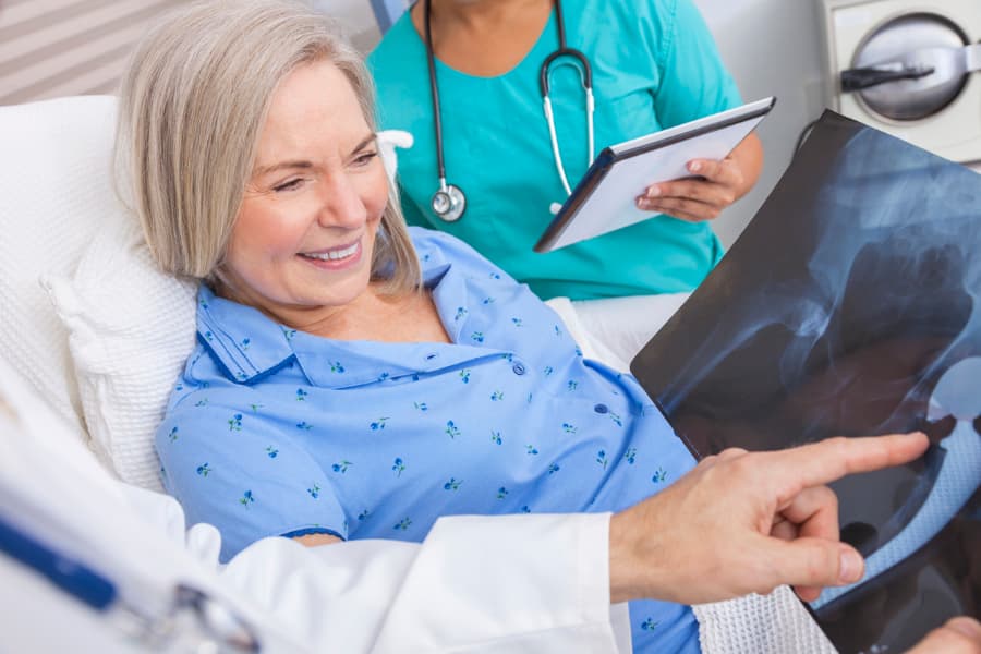 Happy Patient Recovering In Hospital Bed With Physician And Nurse Looking At Hip Replacement X-Ray