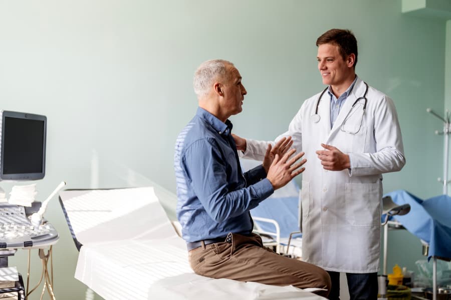 Older male patient sitting on exam table in physician’s office while speaking to urologist 